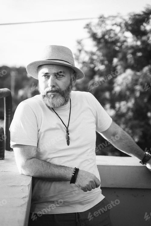 Preview: Adult male in his middle age stands on a balcony wearing a classic fedora hat with a beard
