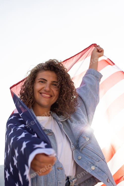 Preview: Happy Beautiful diverse woman holding United States of America flag and standing against sunlight