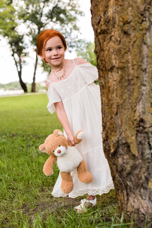 Preview: adorable redhead girl in white dress holding teddy bear and looking away in park