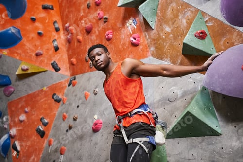Preview: athletic african american male model with alpine harness posing next to rock climbing wall