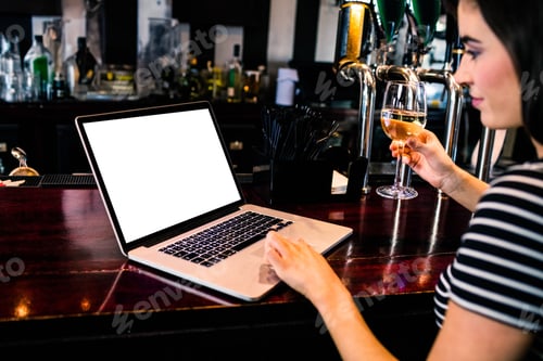 Preview: Woman holding glass of white wine and typing on open laptop at polished wood bar counter
