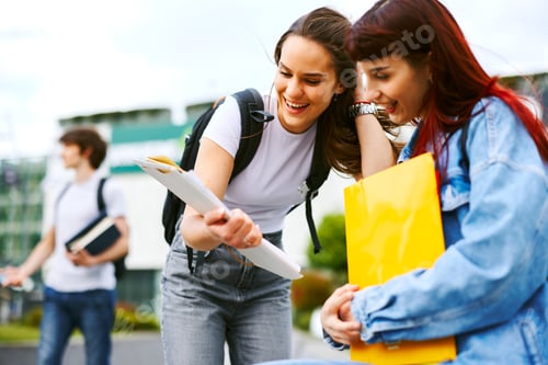 Preview: Two happy female students sharing notes and laughing together on a college campus