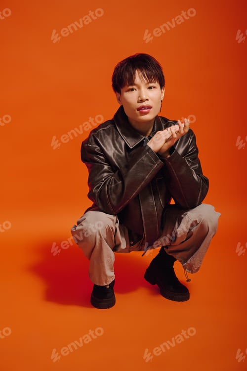 Preview: Stylish young man in trendy attire poses against vibrant orange backdrop