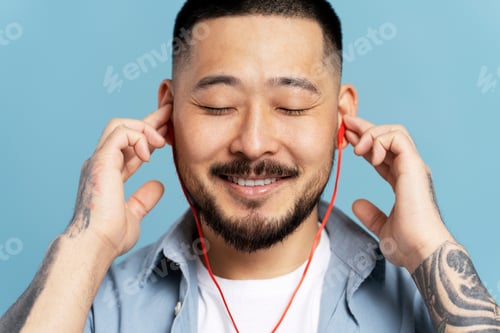 Preview: Smiling asian man with eyes closed, wearing red headphones listening music isolated on blue