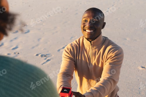Preview: Happy african american man kneeling with ring and proposing to girlfriend on sunny beach