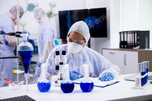 Preview: Female scientist in protection equipment taking notes with smoking blue solution