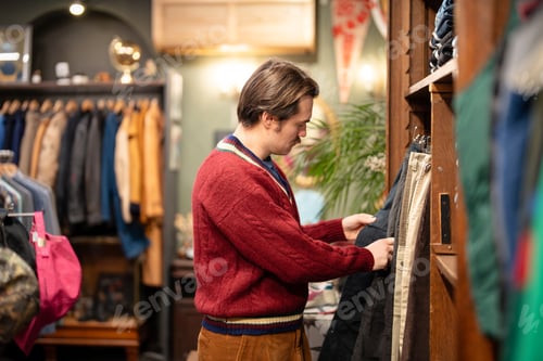 Preview: Attractive man in knit sweater on shopping day in vintage clothing store, buying second hand pants.
