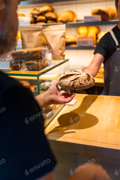 Preview: Hands of customer in the bakery shop buying an artisan bread from the baker in the workshop