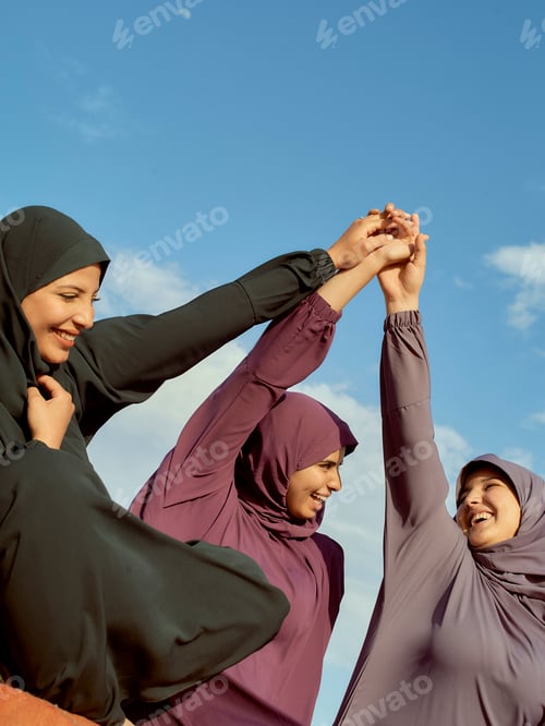 Preview: Three women in hijabs holding hands up together in solidarity under a blue sky.