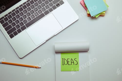 Preview: top view of paper sticker with word ideas in notebook near laptop on white tabletop