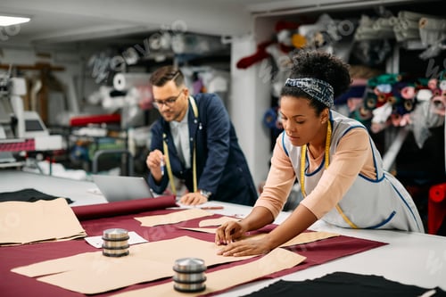 Preview: Young black seamstress using chalk and paper pattern on fabric while working at tailor's.