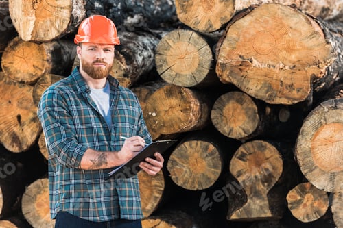 Preview: lumberjack with tattooed hand in protective helmet writing in clipboard on logs background