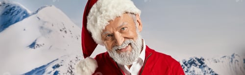 Preview: cheerful white bearded Santa smiling at camera with snowy mountain backdrop, winter concept, banner