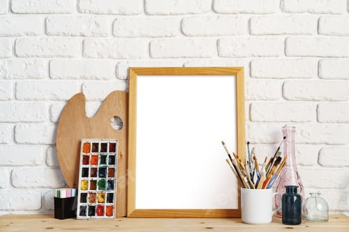 Preview: Photo frame as easel with artist's tools on wooden table against white brick wall