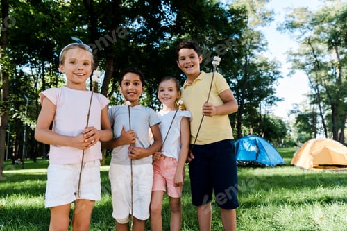 Preview: happy multicultural kids holding sticks with marshmallows near camps