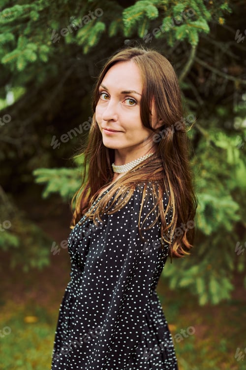 Preview: Young woman with long brown hair gazing amidst lush green pine trees