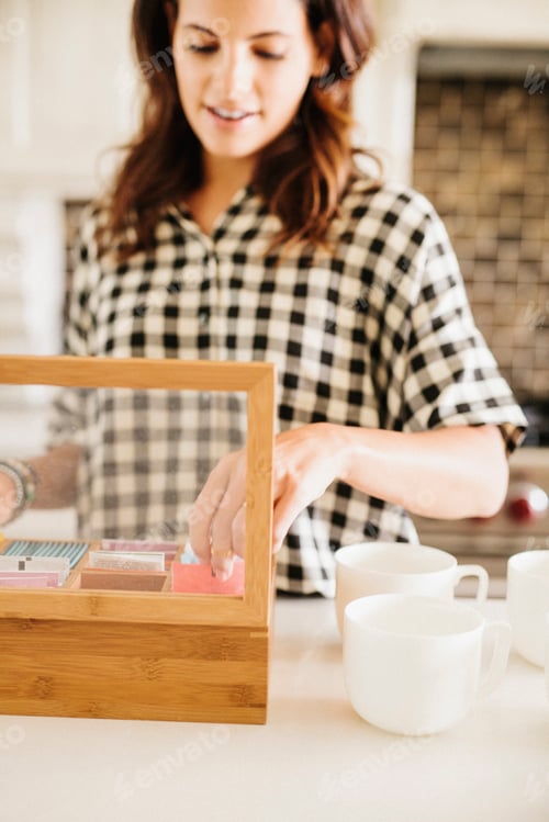 Preview: Woman with long brown hair, wearing a chequered shirt, standing in a kitchen, making a cup of tea.