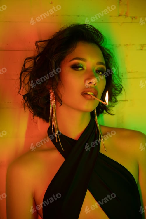 Preview: Portrait of trendy asian woman looking at camera while holding match with fire in lips in night club