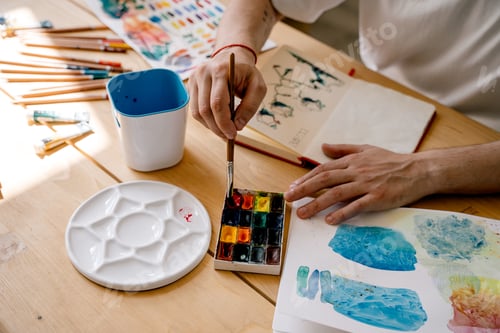 Preview: A young artist paints with watercolors in a notebook sitting at a wooden table. Beautiful male hands