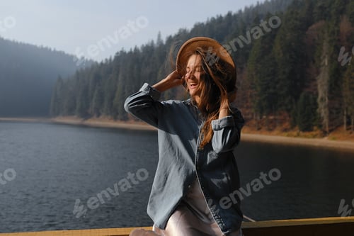 Preview: happy young woman in hat enjoys lake view in forest. Relaxing moments. View of stylish girl enjoys