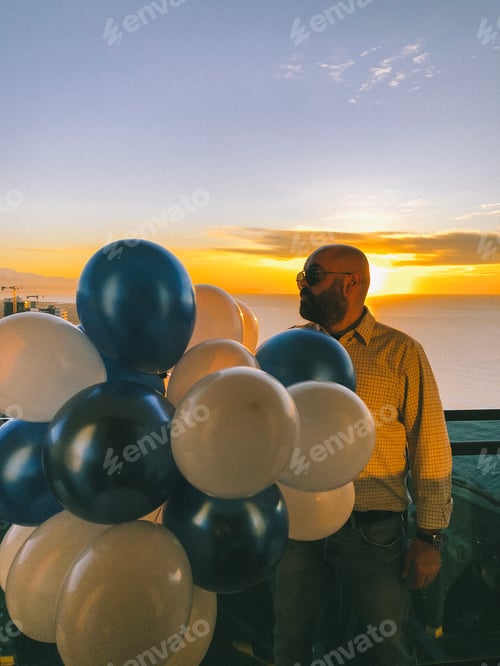 Preview: serious man with a bunch of balloons on the balcony at sunset