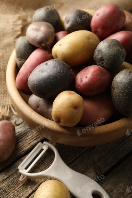 Preview: Different types of potatoes in bowl and peeler on wooden table, closeup