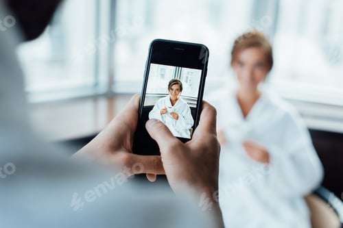 Preview: selective focus of man taking photo of attractive girlfriend holding cup while sitting in bathrobe