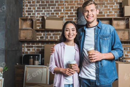 Preview: young interracial couple with paper cups of coffee standing on kitchen while moving into new home