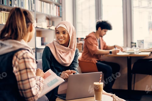 Preview: Happy Muslim woman talking to female friend while studying together in a library.