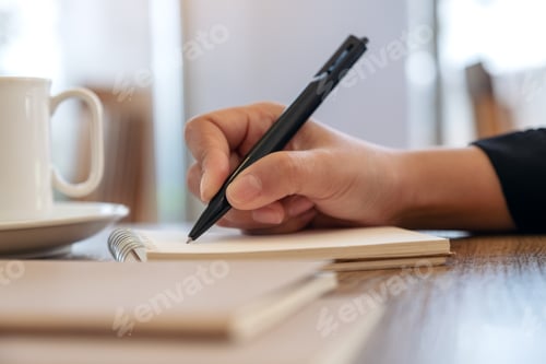Preview: Closeup image of a hand writing on blank notebook with coffee cup on table in cafe
