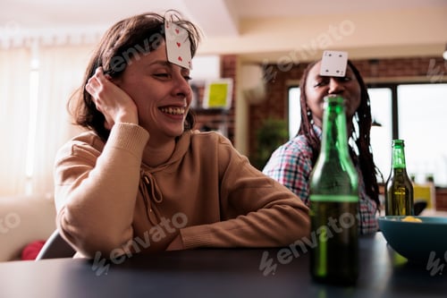 Preview: Smiling woman with card game on forehead enjoying society games with friends at home.