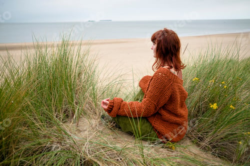 Preview: Woman sitting in sand dunes by the sea, gazing at the horizon on an overcast day, Belgium
