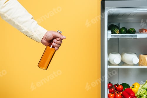 Preview: cropped view of man holding bottle of beer near open fridge with fresh food on shelves isolated on