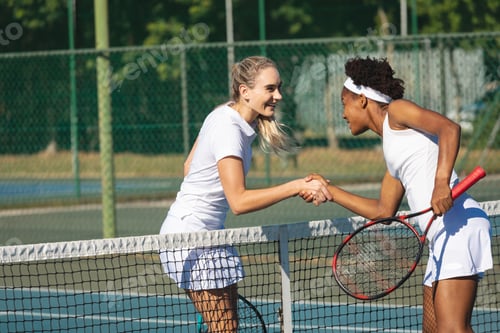 Preview: Happy biracial young female tennis competitors shaking hands over net at court on sunny day