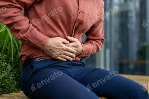 Preview: Businessman outdoors experiencing abdominal pain while sitting on a bench in an urban environment