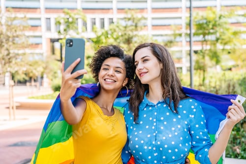 Preview: Smiling multiethnic lesbian couple with rainbow flag taking selfie