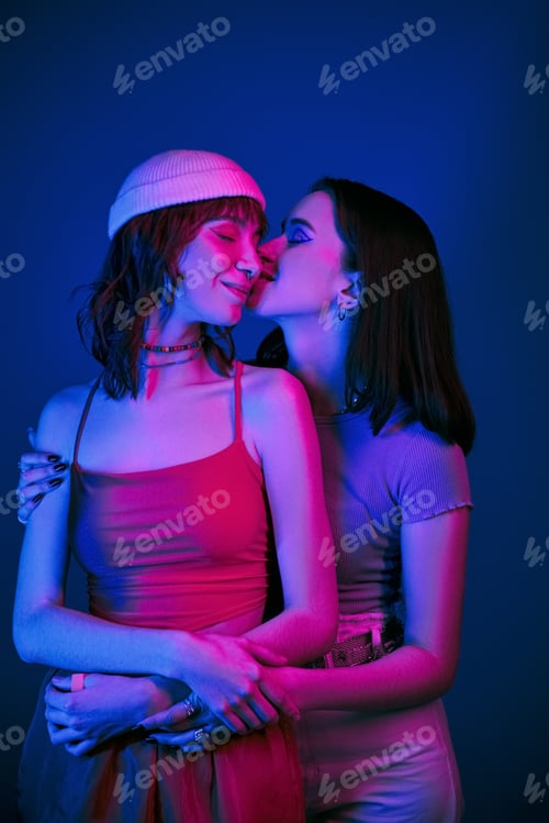Preview: sensual kiss in cheek under purple lights of happy lesbian couple with bold makeup, bliss and love