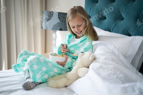 Preview: Smiling girl feeding breakfast to teady bear on bed