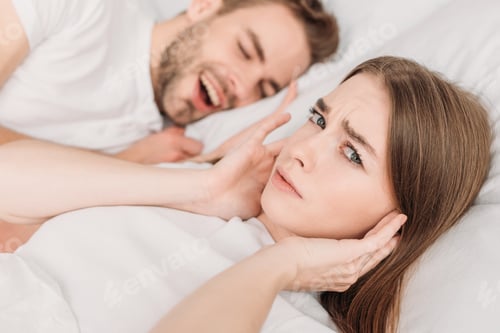 Preview: dissatisfied woman plugging ears with hands and looking at camera while lying in bed near snoring