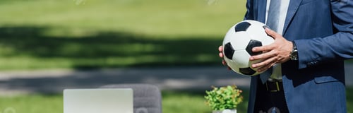 Preview: panoramic shot of businessman holding soccer ball in park near table with laptop