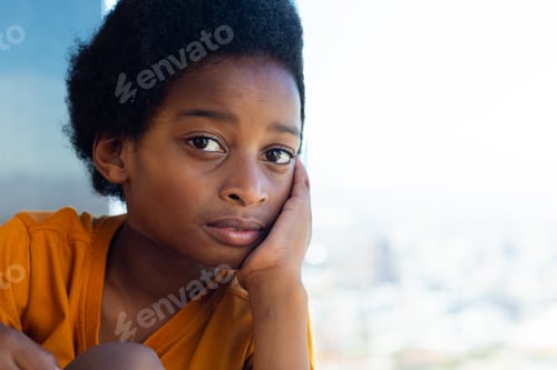 Preview: Sad biracial schoolboy wearing yellow tshirt looking through window in classroom, copy space