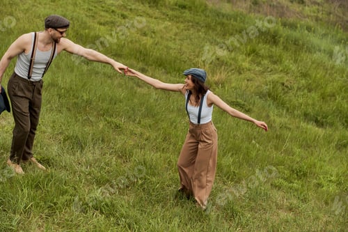 Preview: Side view of positive and fashionable couple in newsboy caps and suspenders holding hands