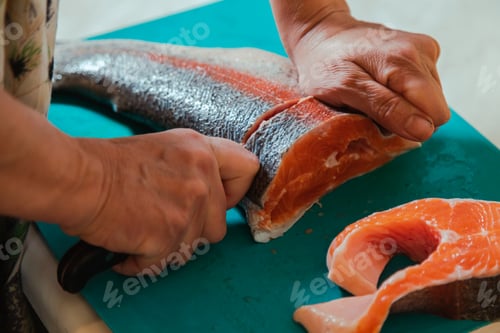 Preview: Hand cutting raw headless salmon fish laying on blue cutting board at home