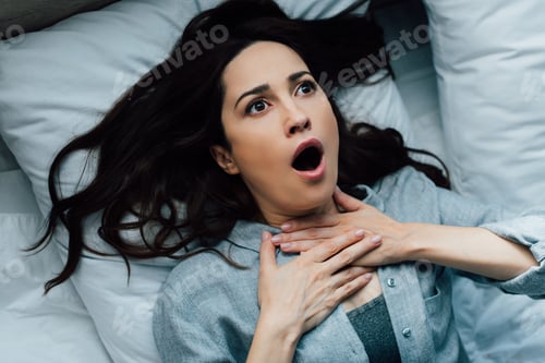 Preview: top view of scared brunette woman touching throat while lying on bed