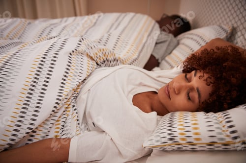 Preview: Millennial African American woman lying on her back asleep in bed, her partner in the background