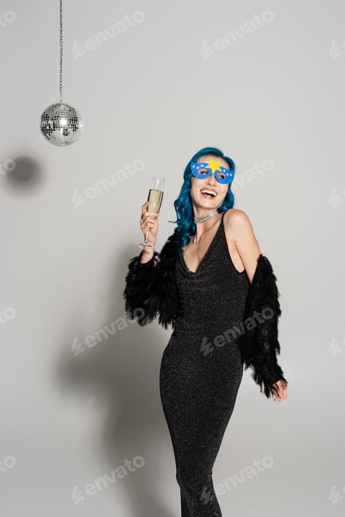 Preview: laughing woman in blue wig and party mask holding champagne and looking away on grey background