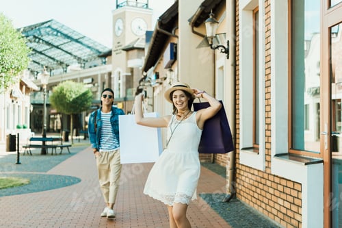 Preview: happy stylish girl with shopping bags walking on the street, her boyfriend walking behind