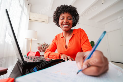 Preview: Cheerful young woman engaged in writing notes while studying with a laptop at home