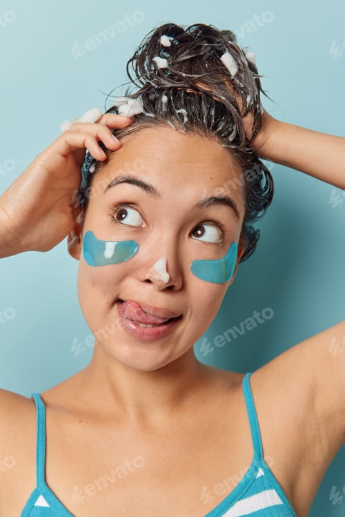 Preview: Vertical shot of thoughtful Asian woman washes hair with shampoo sticks out tongue focused aside dre