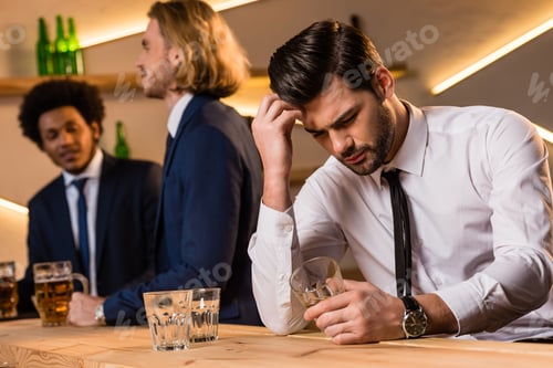 Preview: young drunk businessman sitting at bar counter while coworkers standing behind